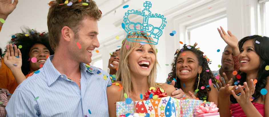 Eine junge Frau mit einem Geschenk in der Hand feiert mit Freunden Geburtstag. Auf dem Foto ist eine gezeichnete Krone auf dem Kopf.