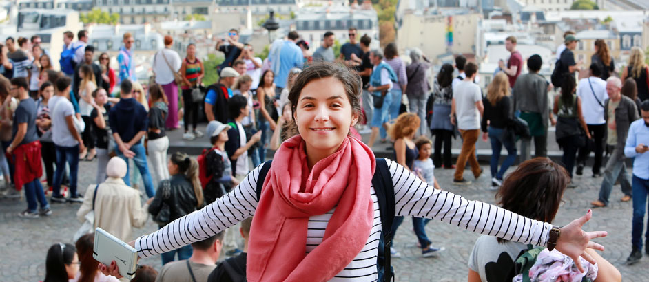 Mädchen steht mit ausgebreiteten Armen vor einer Straße und schaut in die Kamera, auf der Straße hinter ihr gehen viele Menschen vorbei.