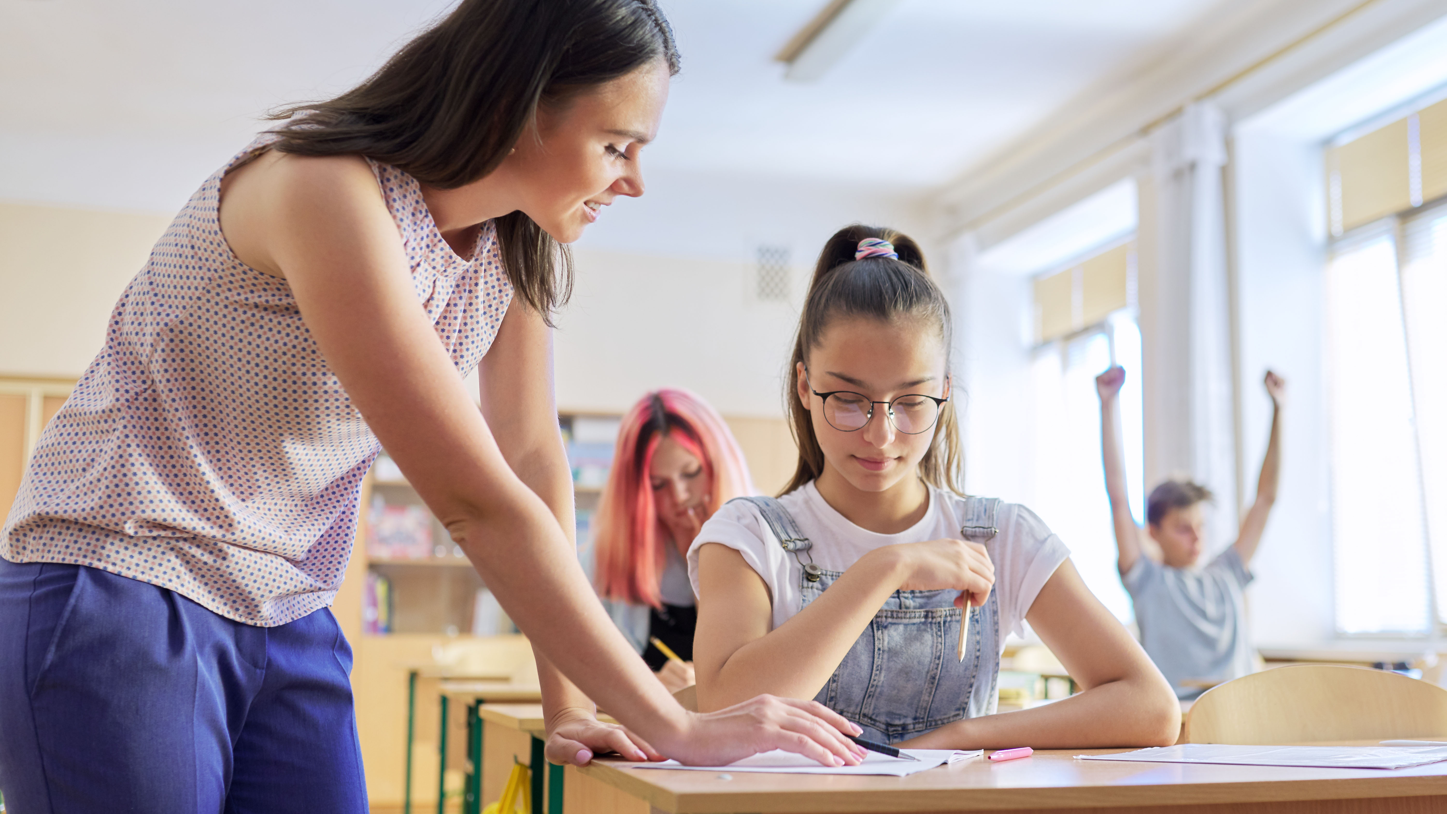 Lehrerin zeigt etwas einer Schülerin, die an ihrem Tisch im Klassenzimmer sitzt.