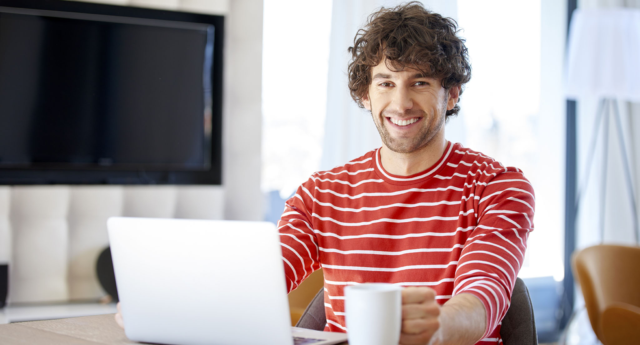 Junger Mann sitzt mit einer Kaffeetasse vor dem Laptop und lächelt in die Kamera