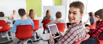Schüler mit Tablet in der Hand sitzt im Klassenzimmer mit seinen Mitschülern und lächelt in die Kamera.
