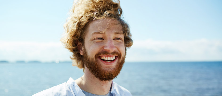 Der Kopf eines lachenden jungen Mannes ist zu sehen, im Hintergrund sieht man das Meer.