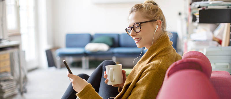 Eine Frau sitzt mit einer Tasse in der Hand auf einer Couch und liest etwas auf ihrem Smartphone.