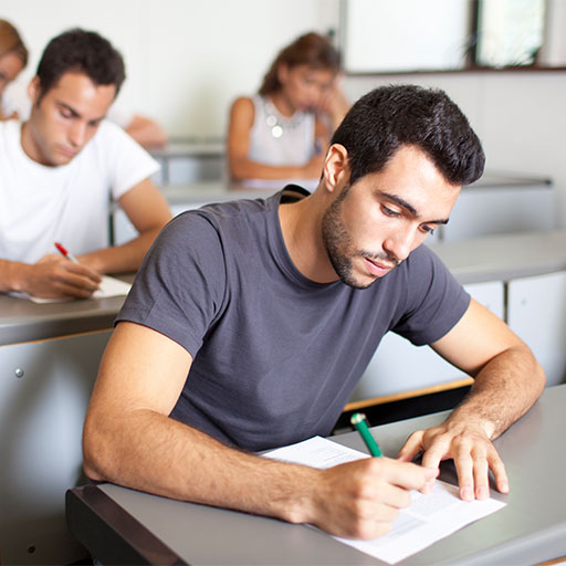 Ein junger Mann im grauen T-Shirt sitzt in einem Kursraum am Tisch und schreibt etwas auf ein Blatt Papier.