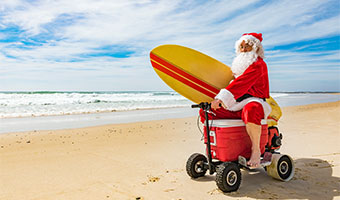 Ein Weihnachtsmann fährt in Australien mit einem Surfboard unterm Arm am Strand entlang. Er sitzt dabei auf einer Kühlbox.