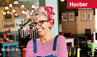 Ältere Frau mit rotweißem Ringelshirt und 50er Jahre Frisur steht lachend in einem Cafe.