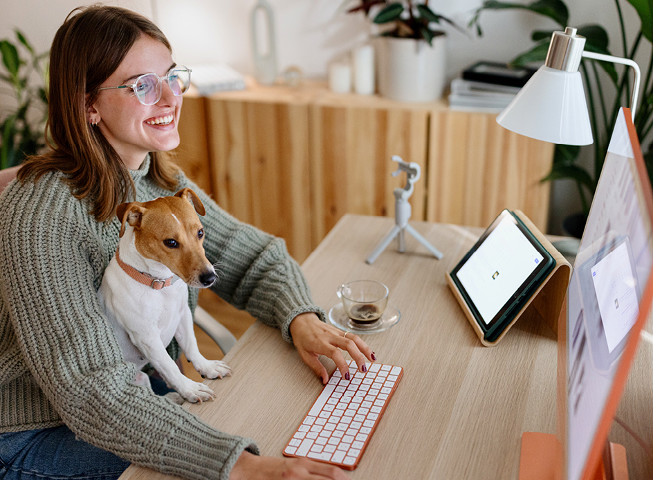 Junge Frau sitzt mit kleinem Hund auf dem Schoss am Schreibtisch und arbeitet am Computer.
