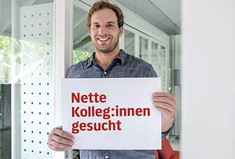 Geschäftsmann lächelt in die Kamera und hält ein Schild mit der Aufschrift
Nette Kolleg:innen gesucht!